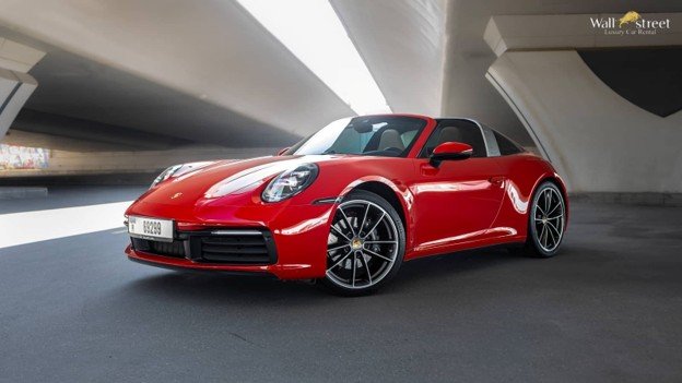 Front three-quarter view of a vibrant red Porsche 911 Targa (992 generation) parked under a modern concrete bridge structure in Dubai, showcasing its distinctive silver Targa bar, open roof, sleek design, and sports car stance with UAE license plates.